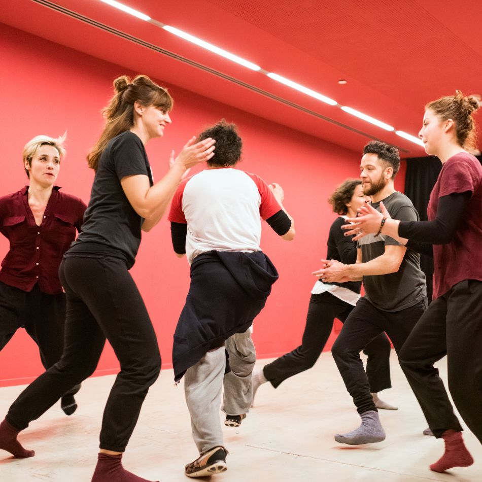 Des personnes participants à un cours de théâtre s'échauffent dans la salle.