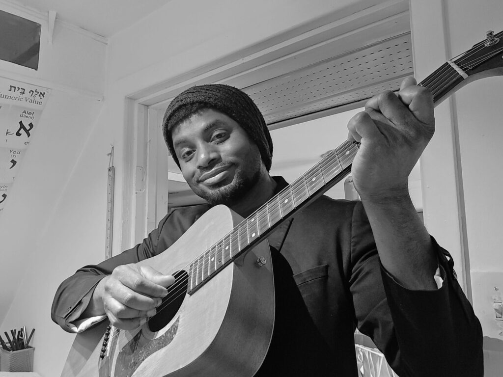Le professeur joue de la guitare en regardant la caméra et en souriant légérement.