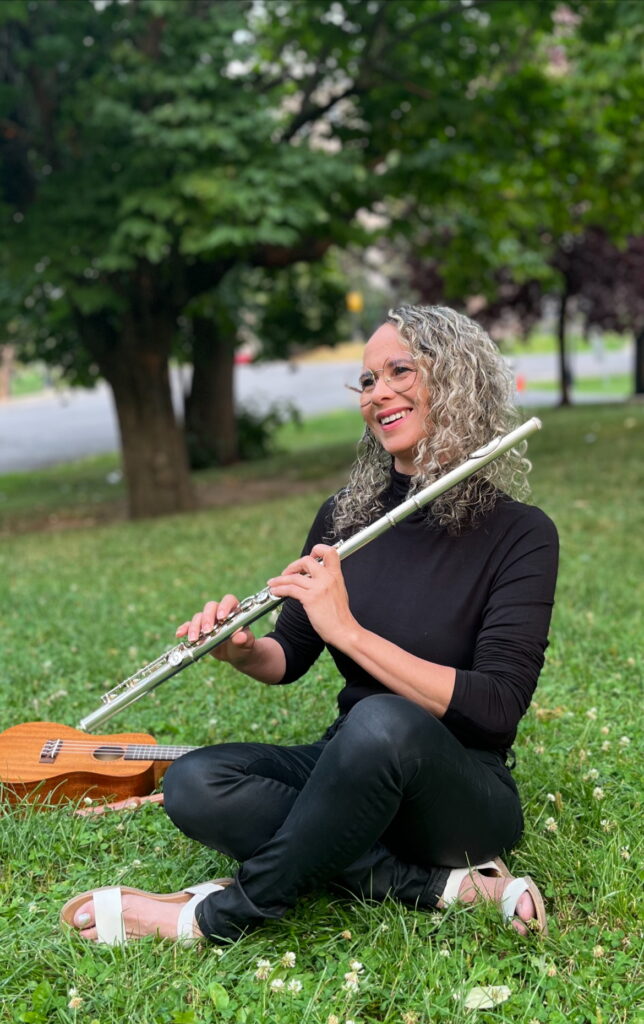 La professeure de musique est assise dans l'herbe. Elle joue de la flûte traversière et une guitare est posée à côté d'elle.